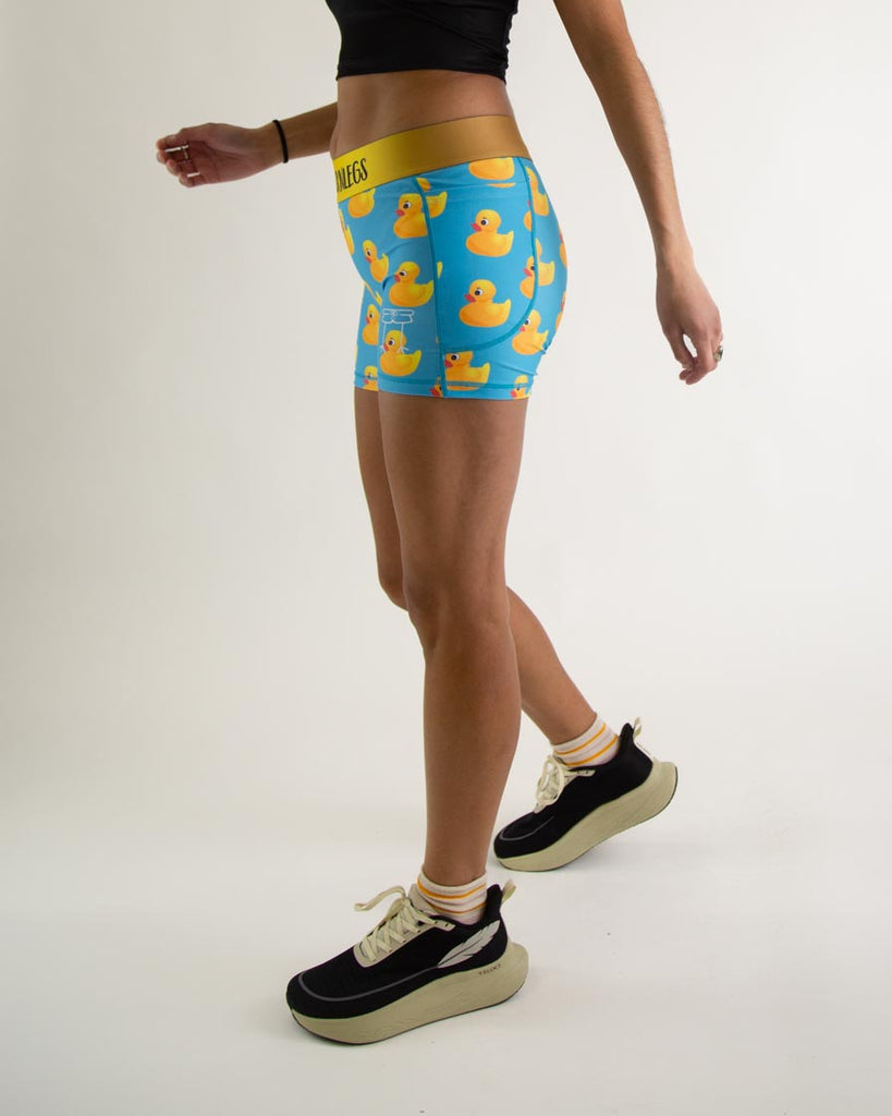 Runner is wearing ChicknLegs in the women's 3 in compression spandex shorts with pockets in the rubber ducky design. The print has yellow rubber ducks on it with a blue background. The model is facing left.