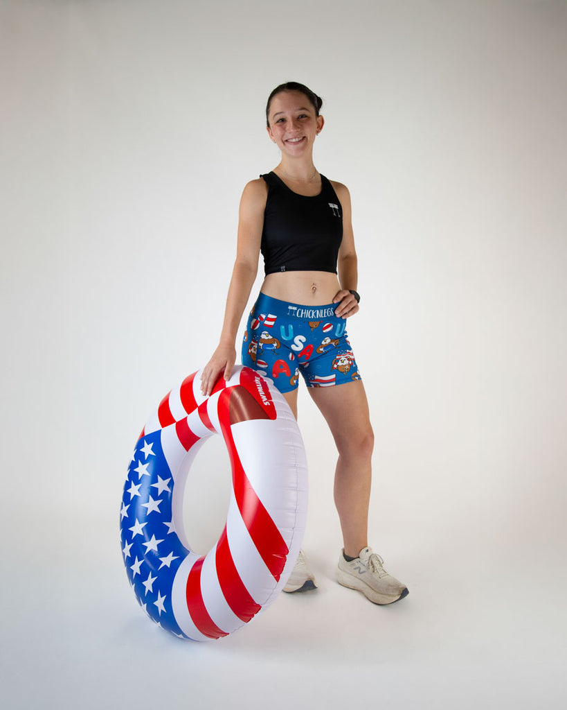 Runner is wearing ChicknLegs in the women's 3 in compression spandex shorts with side pockets. Model is holding a giant pool floatie that has the american flag on it.