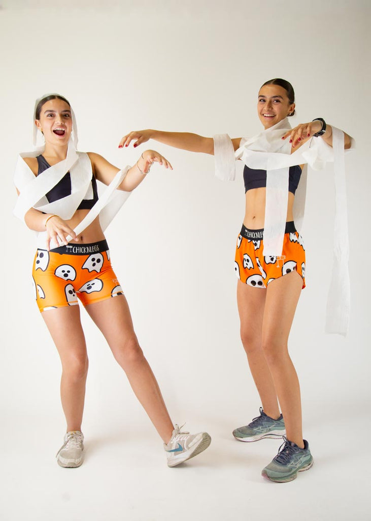 The model is a runner wearing Chicknlegs women's 1.5 inch split running shorts in the Ghosts design, facing front and posing with another model in the women's Chicknlegs 1.5" split running shorts in the Ghost design while posing and wearing toilet paper to be a mummy.
