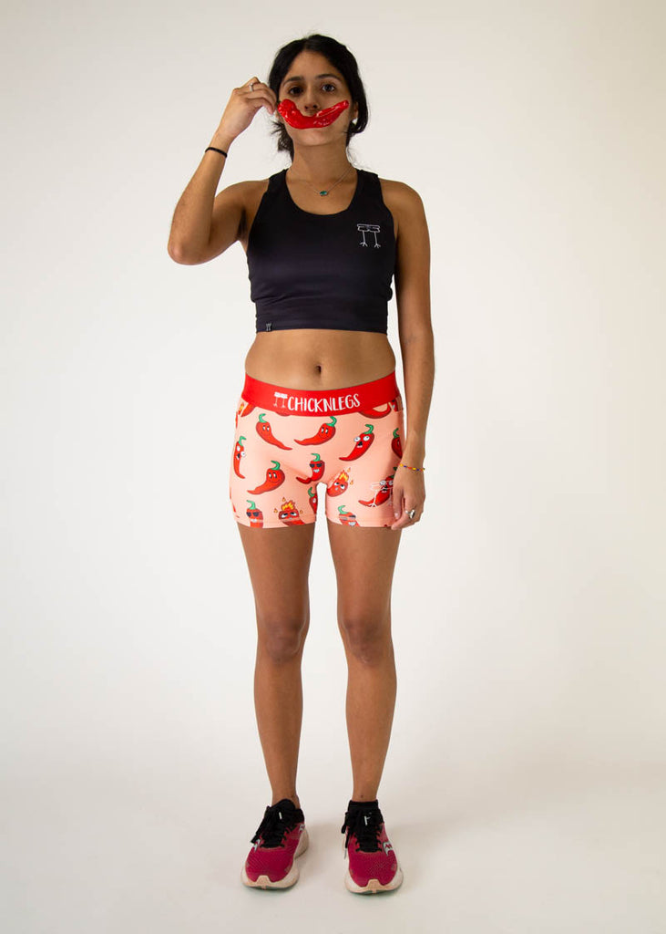 Model is a runner wearing Chicknlegs women's 3 inch compression shorts in the chili pepper design, facing front while posing with a chili pepper in front of their face like it is a smile.