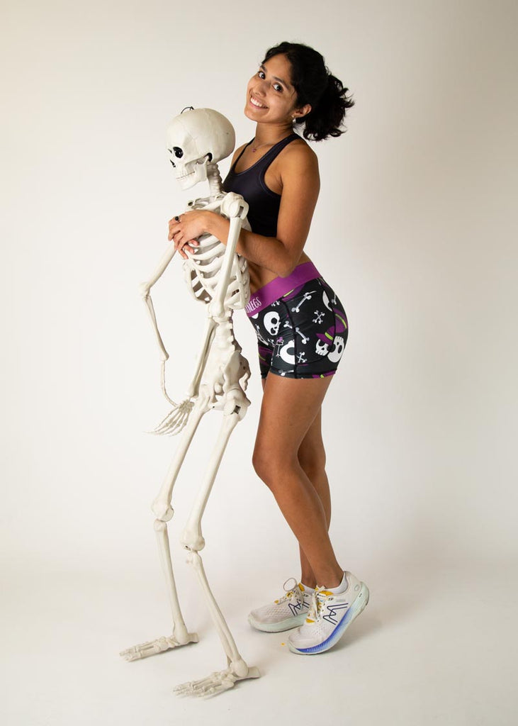 The model is a runner wearing Chicknlegs in women's 3 inch compression shorts in the skulls design, facing front while posing with a skeleton.