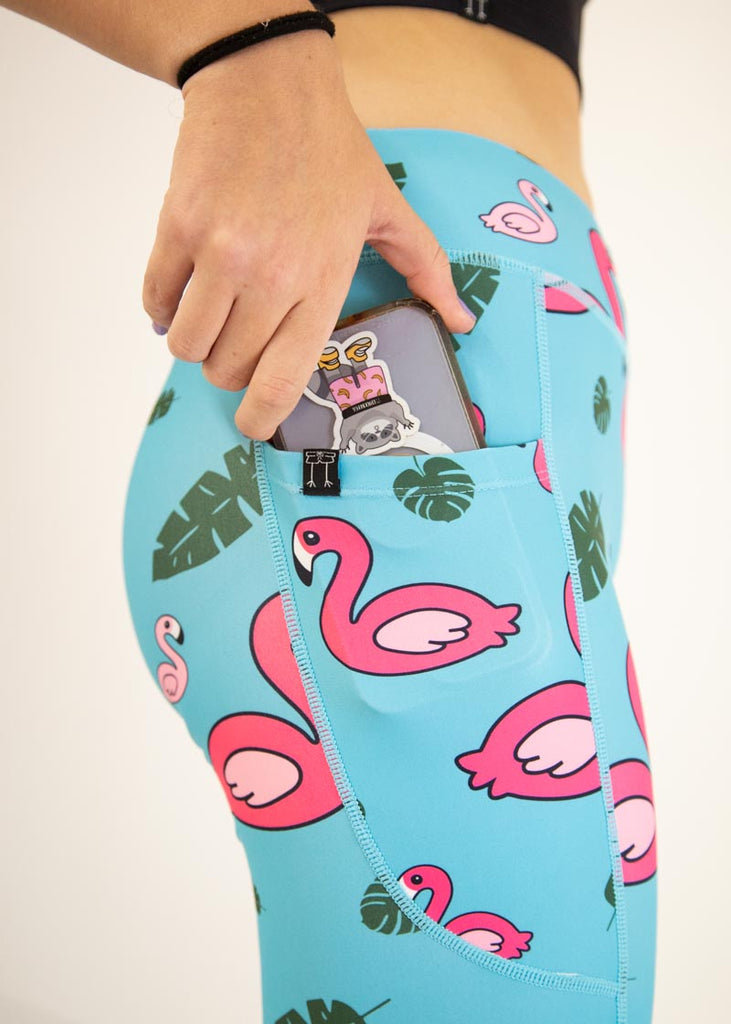 The model is a runner who is wearing Chicknlegs Women's Leggings in the Blue Flamingo design. These leggings include side pockets and an adjustable drawstring at the front. The model is putting a phone into the pocket, a close up is shown.