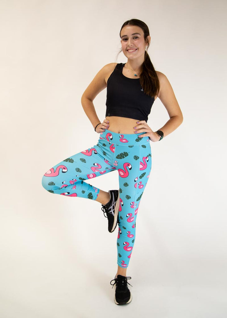 The model is a runner who is wearing Chicknlegs Women's Leggings in the Blue Flamingo design. These leggings include side pockets and an adjustable drawstring at the front. The model is facing front while posing and stretching their leg.