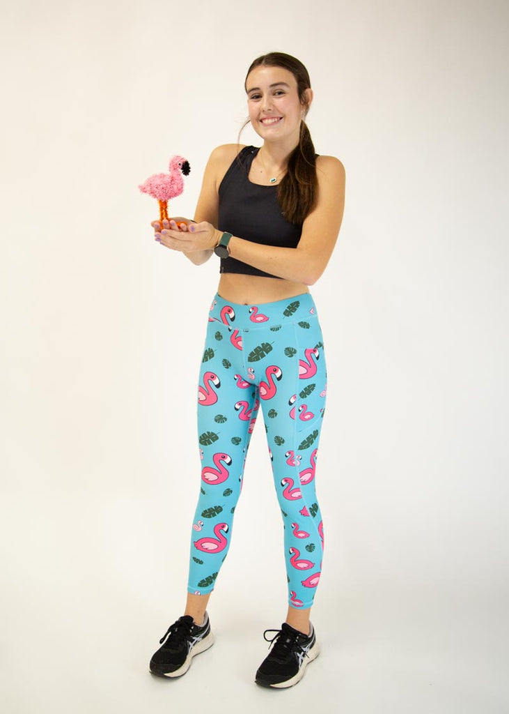 The model is a runner who is wearing Chicknlegs Women's Leggings in the Blue Flamingo design. These leggings include side pockets and an adjustable drawstring at the front. The model is facing front while holding a small flamingo.