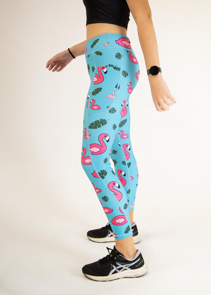 The model is a runner who is wearing Chicknlegs Women's Leggings in the Blue Flamingo design. These leggings include side pockets and an adjustable drawstring at the front. The model is facing left for a side view.