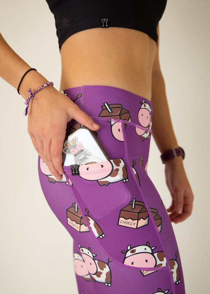 The model is a runner who is wearing Chicknlegs Women's Leggings in the Choccy Cow design. These leggings include side pockets and an adjustable drawstring at the front. The model is putting their phone into the side pocket, a close up image is shown.