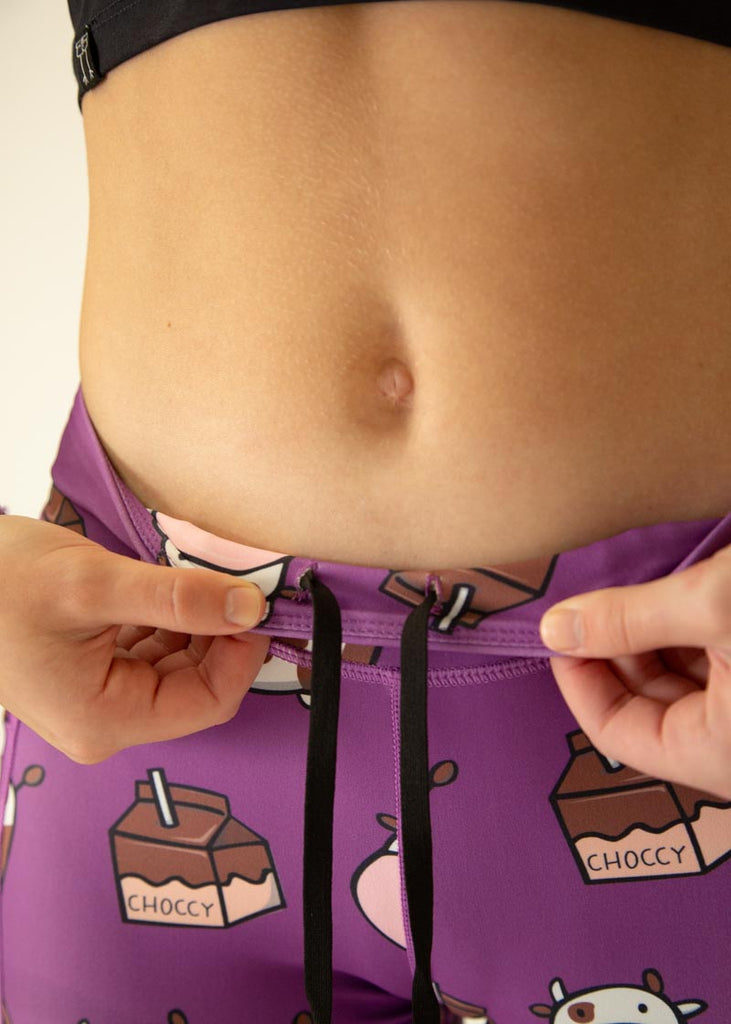 The model is a runner who is wearing Chicknlegs Women's Leggings in the Choccy Cow design. These leggings include side pockets and an adjustable drawstring at the front. The model is facing front while showing the drawstrings.