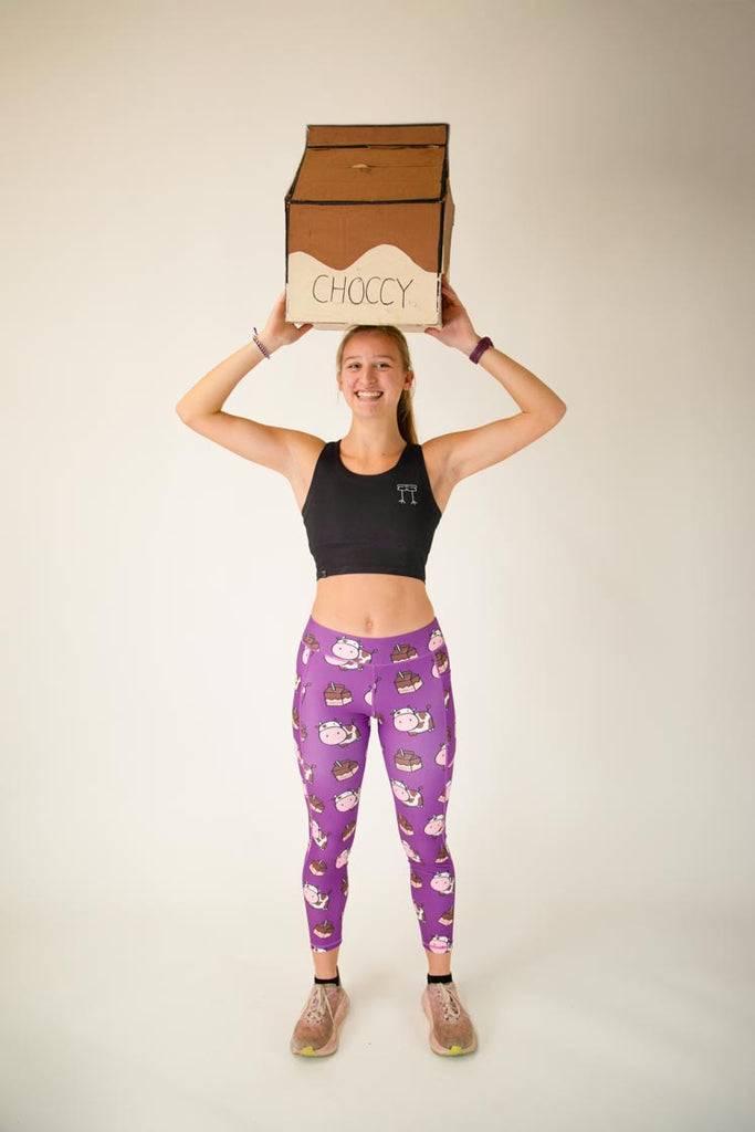 The model is a runner who is wearing Chicknlegs Women's Leggings in the Choccy Cow design. These leggings include side pockets and an adjustable drawstring at the front. The model is facing front while posing with a giant chocolate milk carton.