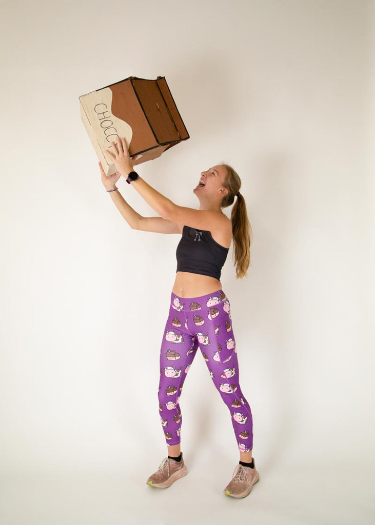 The model is a runner who is wearing Chicknlegs Women's Leggings in the Choccy Cow design. These leggings include side pockets and an adjustable drawstring at the front. The model is posing with a giant chocolate milk carton.
