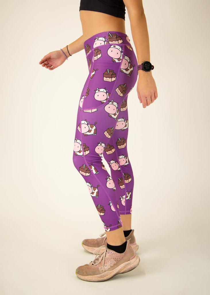 The model is a runner who is wearing Chicknlegs Women's Leggings in the Choccy Cow design. These leggings include side pockets and an adjustable drawstring at the front. The model is facing left for a side view.