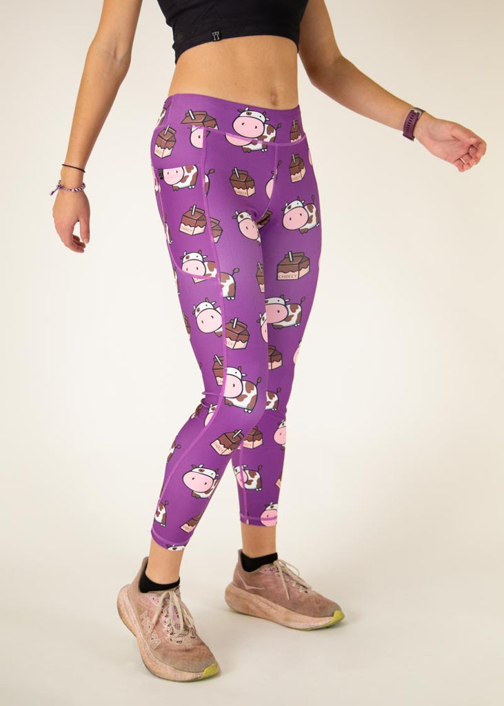 The model is a runner who is wearing Chicknlegs Women's Leggings in the Choccy Cow design. These leggings include side pockets and an adjustable drawstring at the front. The model is facing slightly right to show the leggings.