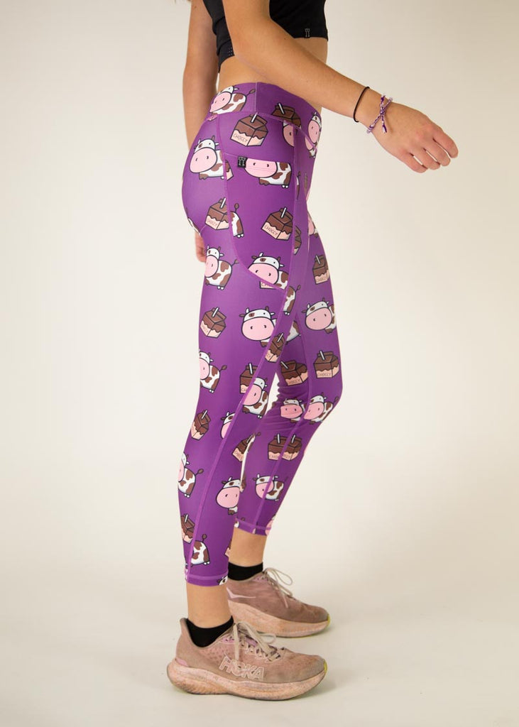 The model is a runner who is wearing Chicknlegs Women's Leggings in the Choccy Cow design. These leggings include side pockets and an adjustable drawstring at the front. The model is facing right to show the side of the leggings.