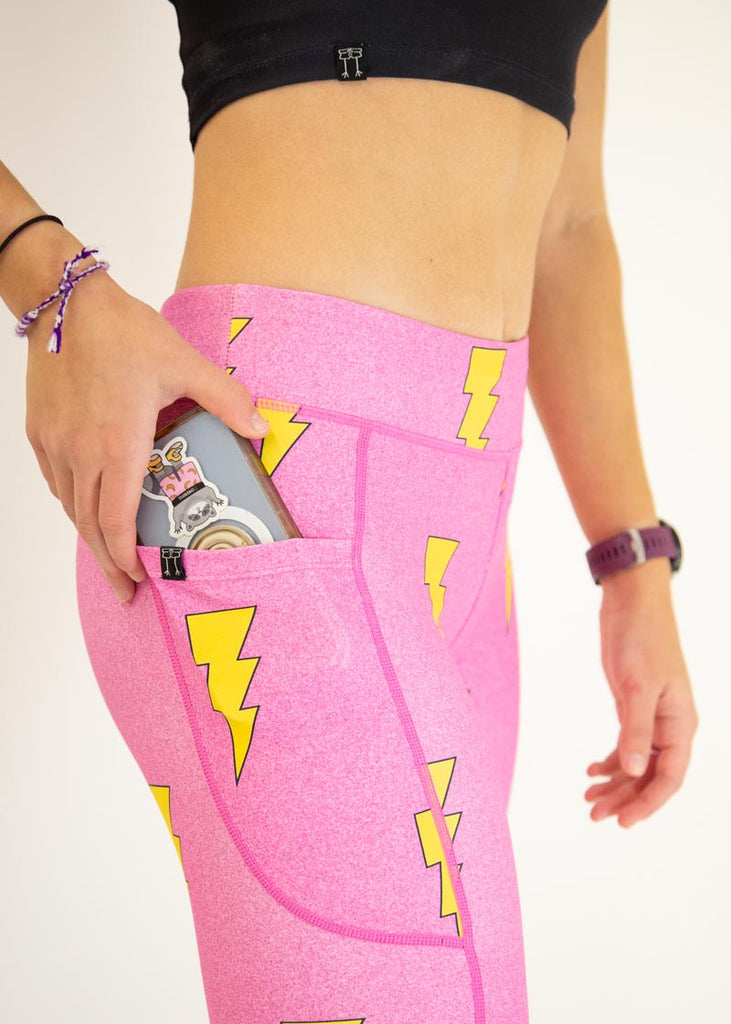 The model is a runner who is wearing Chicknlegs Women's Leggings in the Hot Pink Bolts design. These leggings include side pockets and an adjustable drawstring at the front. The model is facing to the side and putting a phone in their pocket. A close up image is shown.