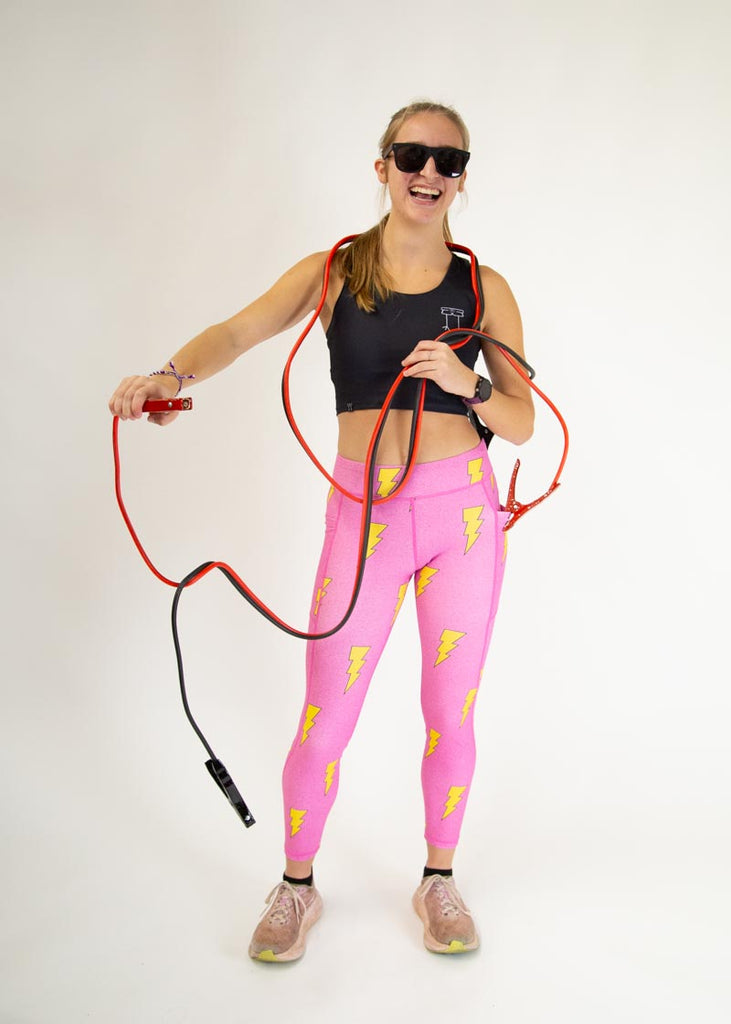 The model is a runner who is wearing Chicknlegs Women's Leggings in the Hot Pink Bolts design. These leggings include side pockets and an adjustable drawstring at the front. The model is facing front while charging themselves with an electrical cord.