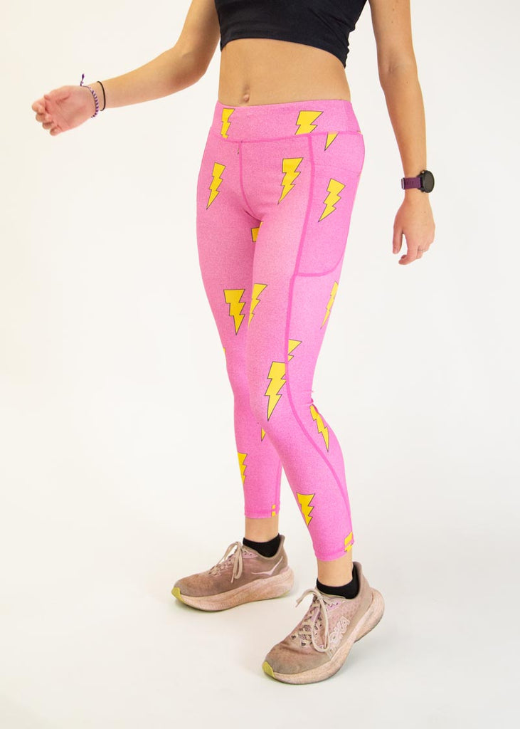 The model is a runner who is wearing Chicknlegs Women's Leggings in the Hot Pink Bolts design. These leggings include side pockets and an adjustable drawstring at the front. The model is facing slightly left to show the side.