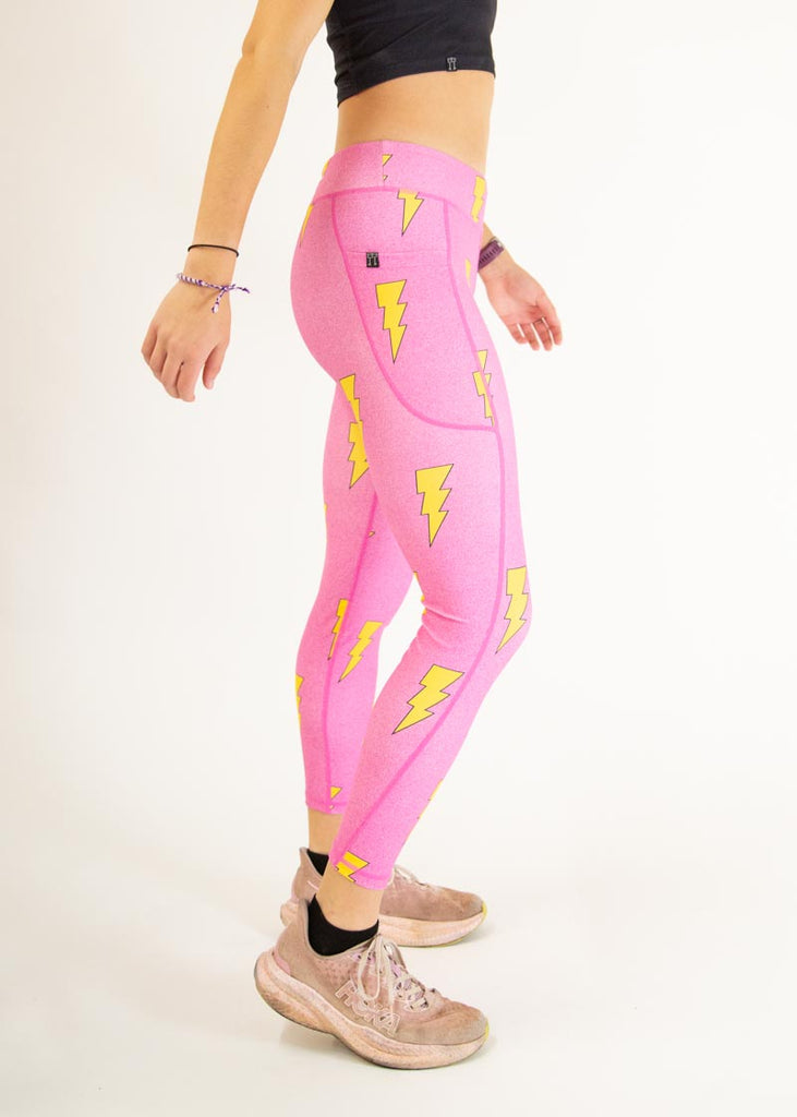 The model is a runner who is wearing Chicknlegs Women's Leggings in the Hot Pink Bolts design. These leggings include side pockets and an adjustable drawstring at the front. The model is facing right to show the side of the pocket.
