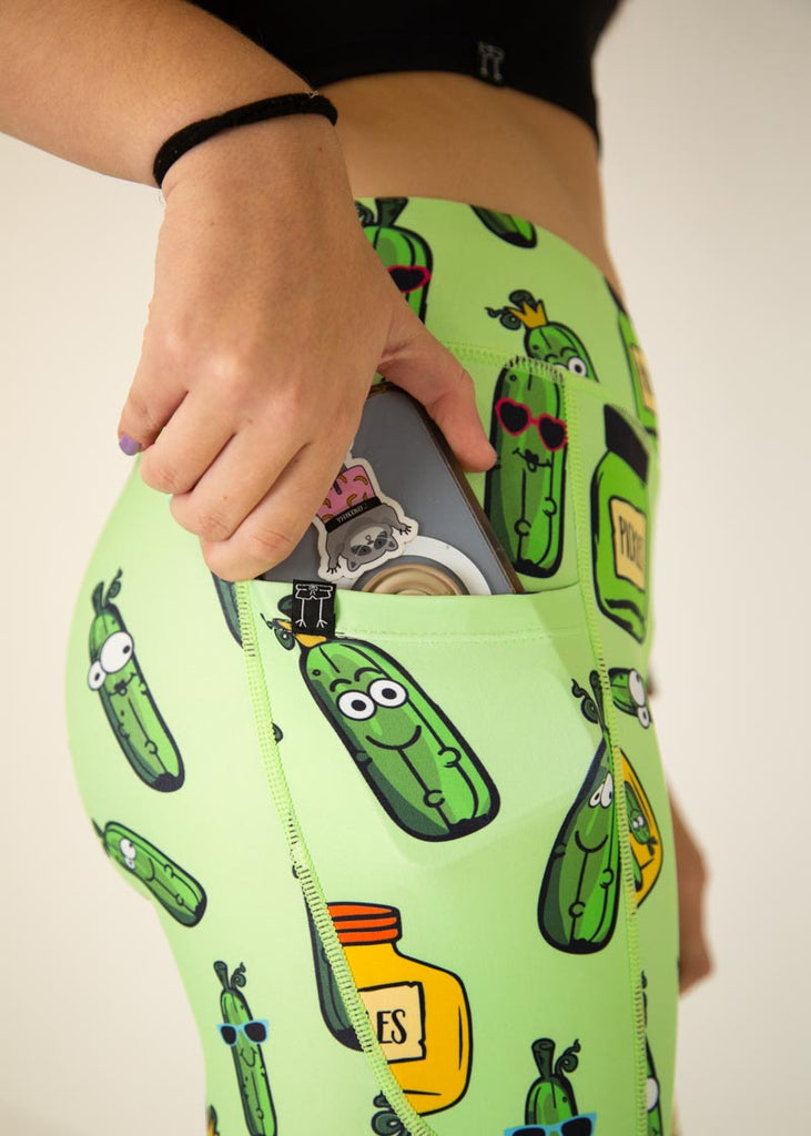 The model is a runner who is wearing Chicknlegs Women's Leggings in the Pickles design. These leggings include side pockets and an adjustable drawstring at the front. The model is facing the right and putting a phone into their pocket.