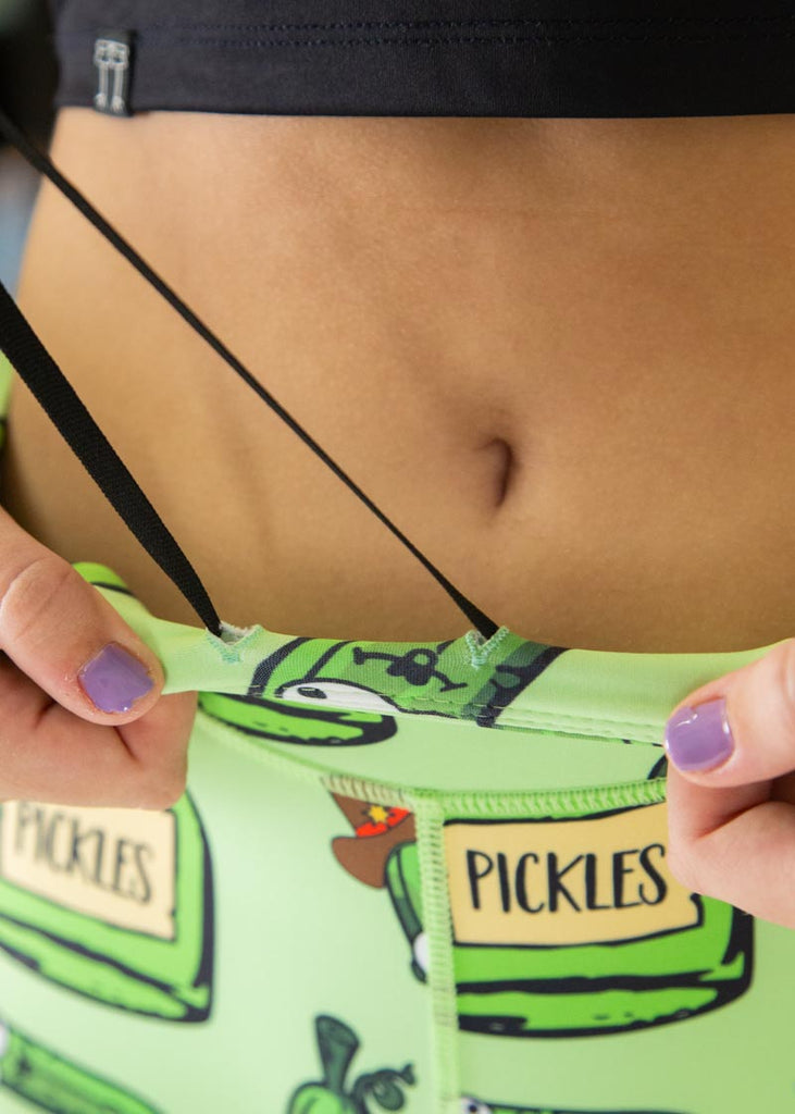 The model is a runner who is wearing Chicknlegs Women's Leggings in the Pickles design. These leggings include side pockets and an adjustable drawstring at the front. The model is facing front while having their drawstrings pulled at.