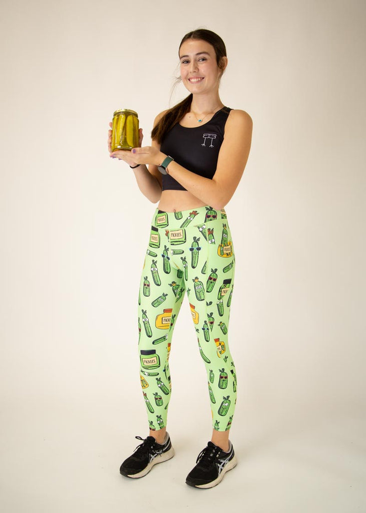 The model is a runner who is wearing Chicknlegs Women's Leggings in the Pickles design. These leggings include side pockets and an adjustable drawstring at the front. The model is facing front while holding a jar of pickles to match the pickle leggings.