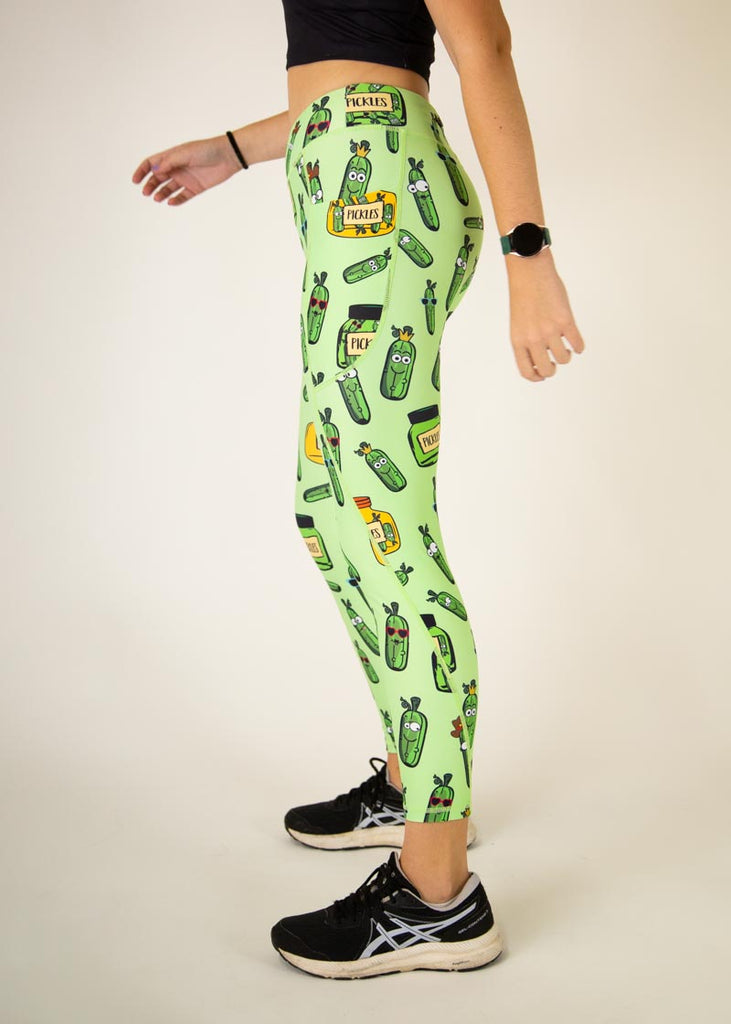 The model is a runner who is wearing Chicknlegs Women's Leggings in the Pickles design. These leggings include side pockets and an adjustable drawstring at the front. The model is facing left to show the side of the pocket.