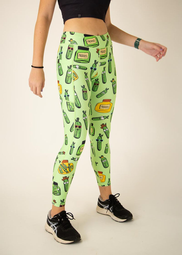 The model is a runner who is wearing Chicknlegs Women's Leggings in the Pickles design. These leggings include side pockets and an adjustable drawstring at the front. The model is facing slightly right to show the side.