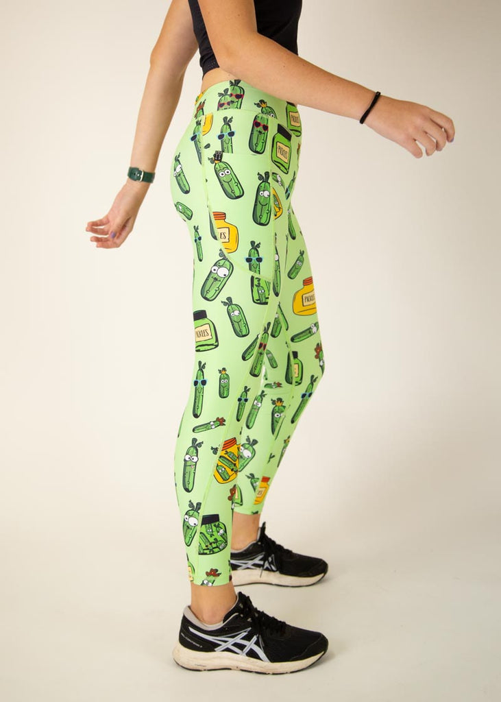 The model is a runner who is wearing Chicknlegs Women's Leggings in the Pickles design. These leggings include side pockets and an adjustable drawstring at the front. The model is facing the right side to show the side of the leggings.