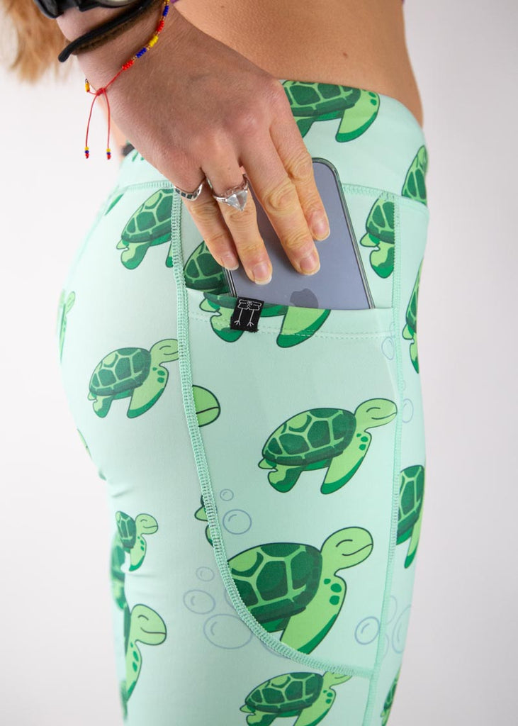 The model is a runner who is wearing Chicknlegs Women's Leggings in the Sea Turtles design. These leggings include side pockets and an adjustable drawstring at the front. The model is facing to the side and putting a phone into their pocket. A close up image is shown.