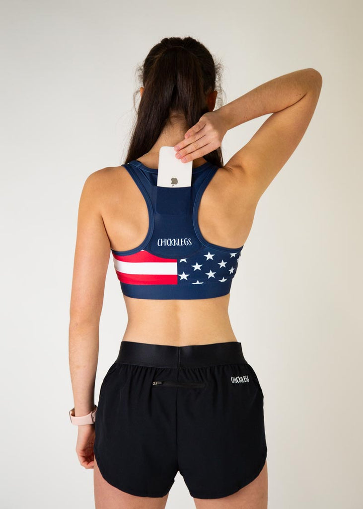 The model is a runner who is wearing Chicknlegs in the women's OG sports bra, in the USA design, facing away from the camera to show the back of the sports bra the model is holding a phone and is putting it into the back pocket.