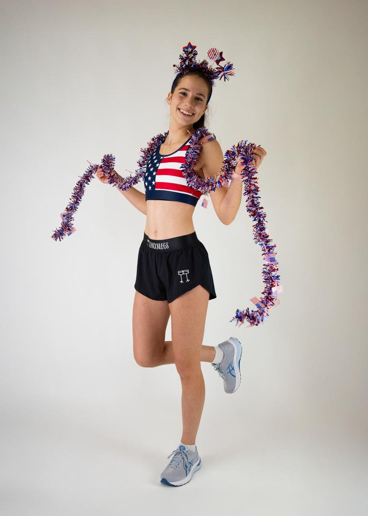 The model is a runner who is wearing Chicknlegs in the women's OG sports bra, in the USA design, facing front for a full body pose and wearing USA theme tinsel as well as a USA theme headband.