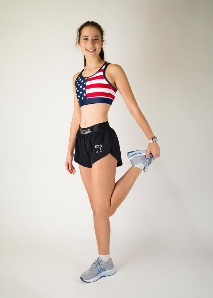 The model is a runner who is wearing Chicknlegs in the women's OG sports bra, in the USA design, facing front for a full body pose while stretching.