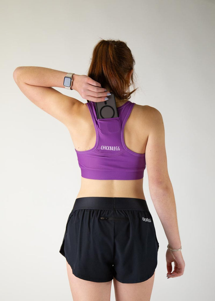 The model is a runner who is wearing Chicknlegs in thhe women's OG sports bra in the deep purple design, facing away from the camera to show the back of the sports bra. The model is holding a phone and putting it into the back of the pocket thats on the sports bra.