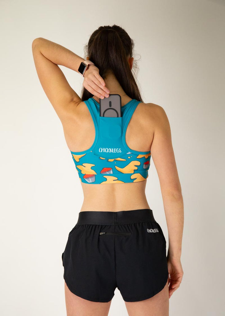 The model is a runner who is wearing Chicknlegs in the OG sports bra, in the dino nuggets design, facing away from the camera to show the back of the sports bra. The model is holding a phone to show it going into the sports bra's pocket.