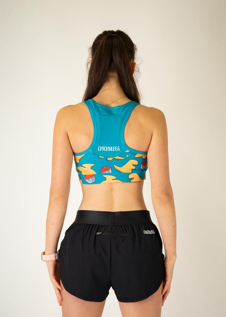The model is a runner who is wearing Chicknlegs in the OG sports bra, in the dino nuggets design, facing away from the camera to show the back of the sports bra.