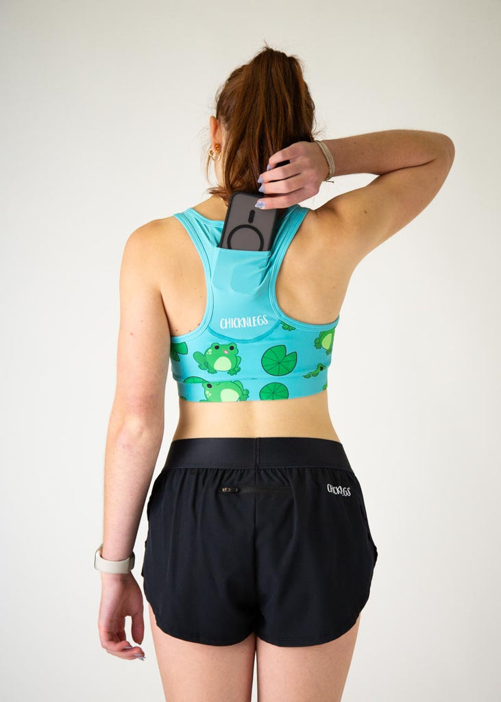 The model is a runner who is wearing Chicknlegs in the women's OG sports bra, in the fartlek froggies frog design, facing away from the camera to show the back of the sports bra while holding a phone to put it into the back pocket.