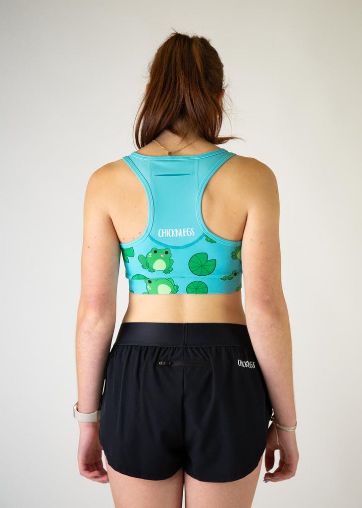 The model is a runner who is wearing Chicknlegs in the women's OG sports bra, in the fartlek froggies frog design, facing away from the camera to show the back of the sports bra.