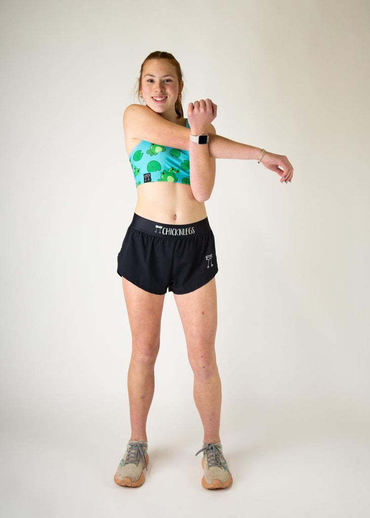 The model is a runner who is wearing Chicknlegs in the women's OG sports bra, in the fartlek froggies frog design, facing front while stretching. A full body image is shown of the model.