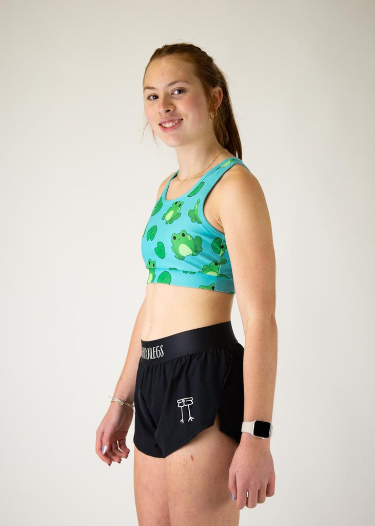 The model is a runner who is wearing Chicknlegs in the women's OG sports bra, in the fartlek froggies frog design, facing left.