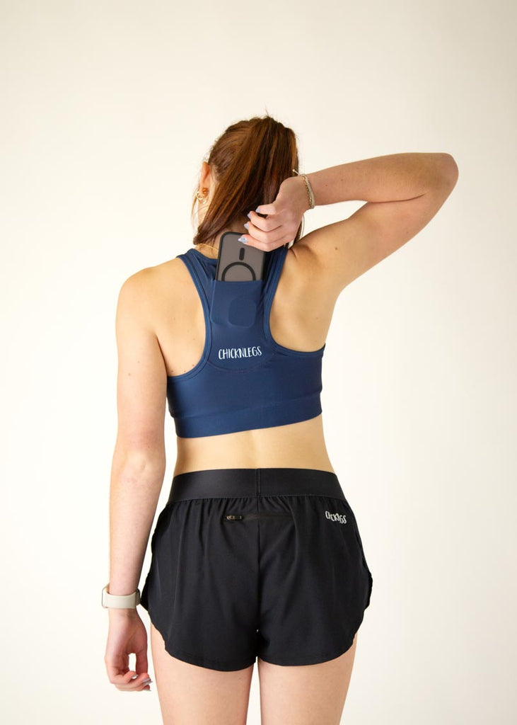 The model is a runner who is wearing Chicknlegs in the women's OG sports bra in the navy design, facing away from the camera to show the back of the sports bra while holding their phone to show it going into the pocket of the bra.
