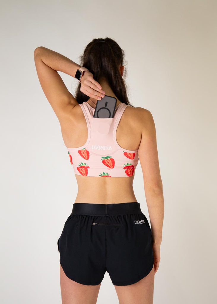 The model is a runner who is wearing Chicknlegs in the women's OG sports bra, in the strawberry SZN design, facing away from the camera to show the back of the sports bra, the model is holding a phone to show it going into the back of the pocket that is on the sports bra.