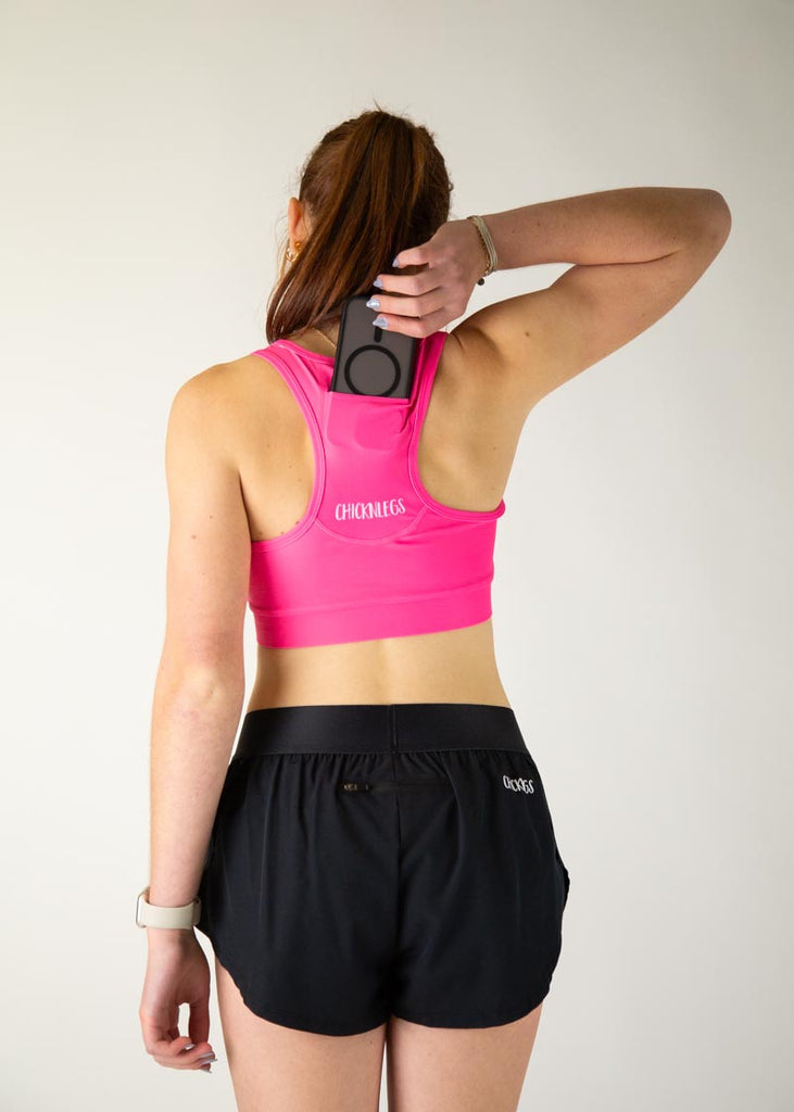 The model is a runner who is wearing Chicknlegs in the women's OG sports bra in the neon pink design, facing away from the camera to show the back of the sports bra while holding a phone in their hand to show the phone going into the pocket on the sports bra.
