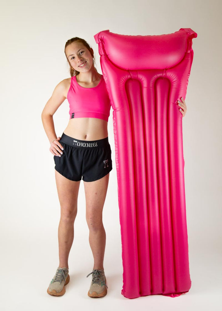 The model is a runner who is wearing Chicknlegs in the women's OG sports bra in the neon pink design, facing front while posing with a huge pink floatie. A full body image is shown of the model.