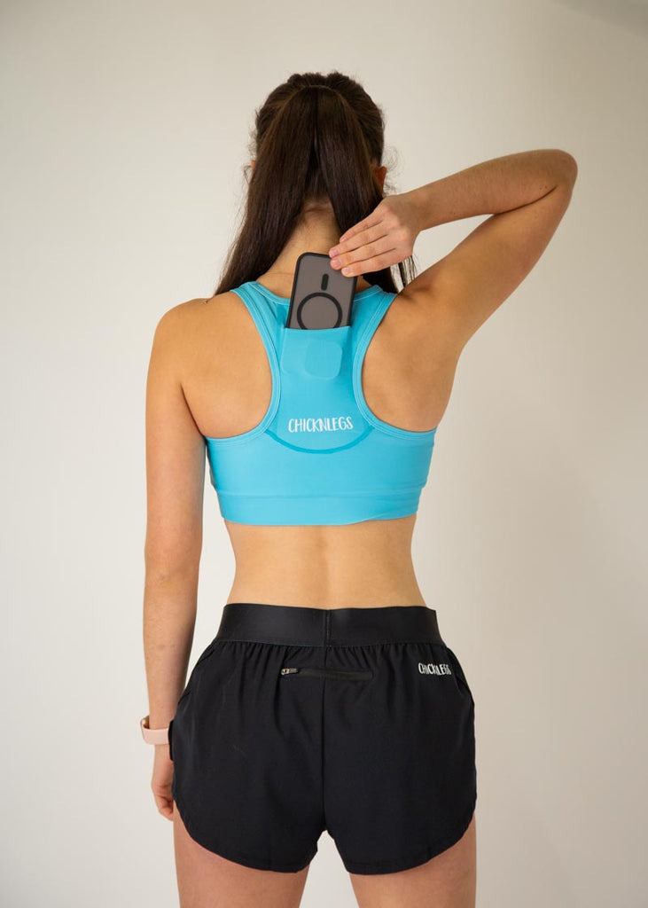 The model is a runner who is wearing Chicknlegs in the women's OG sports bra in the sky blue design, facing away from the camera to show the back of the sports bra. The model is holding a phone and putting it into the sports bra pocket. 