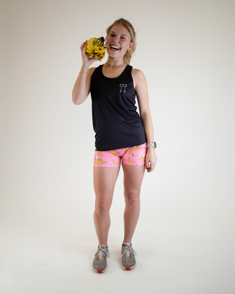 Runner is wearing ChicknLegs in the women's performance singlet in the solid black design and pink banana compression shorts while holding bananas.