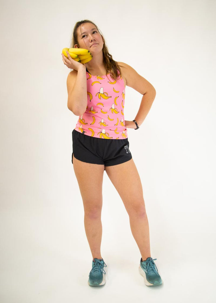 The model is a runner who is wearing Chicknlegs women's performance singlet in the pink banana design, facing front while posing with a banana.