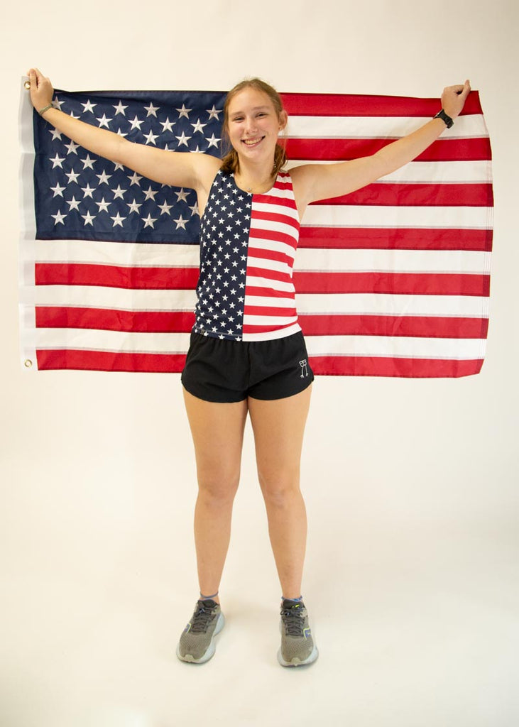 The model is a runner who is wearing Chicknlegs women's singlet in the USA design, facing front while posing with the american flag.