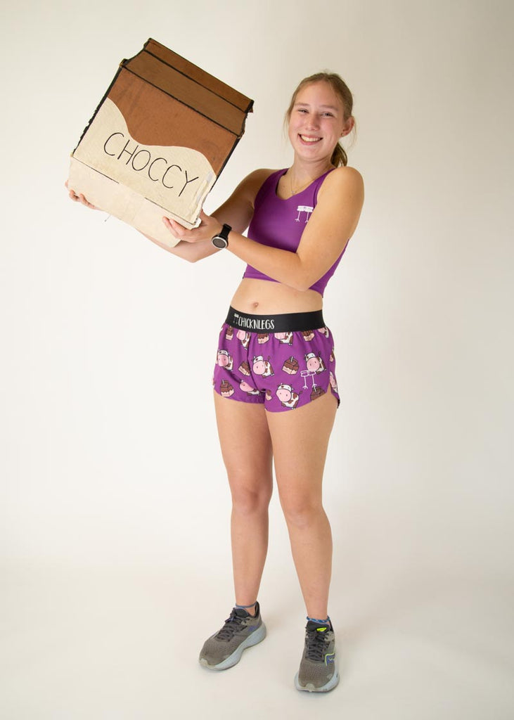 The model is a runner who is wearing Chicknlegs in women's pwr crop top in the deep purple design, matching with the choccy cow women's 1.5" split running shorts. The model is posing with a giant chocolate milk carton.