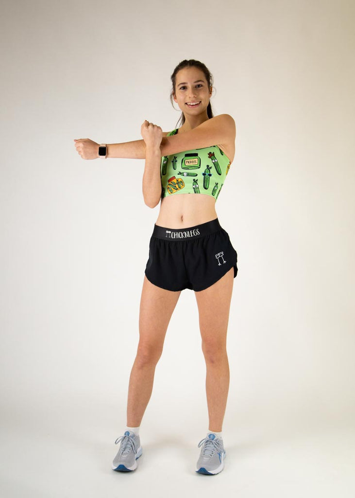 The model is a runner who is wearing Chicknlegs in the women's PWR crop top, in the pickles design, facing front while posing and stretching their arms. A full body photo is shown.