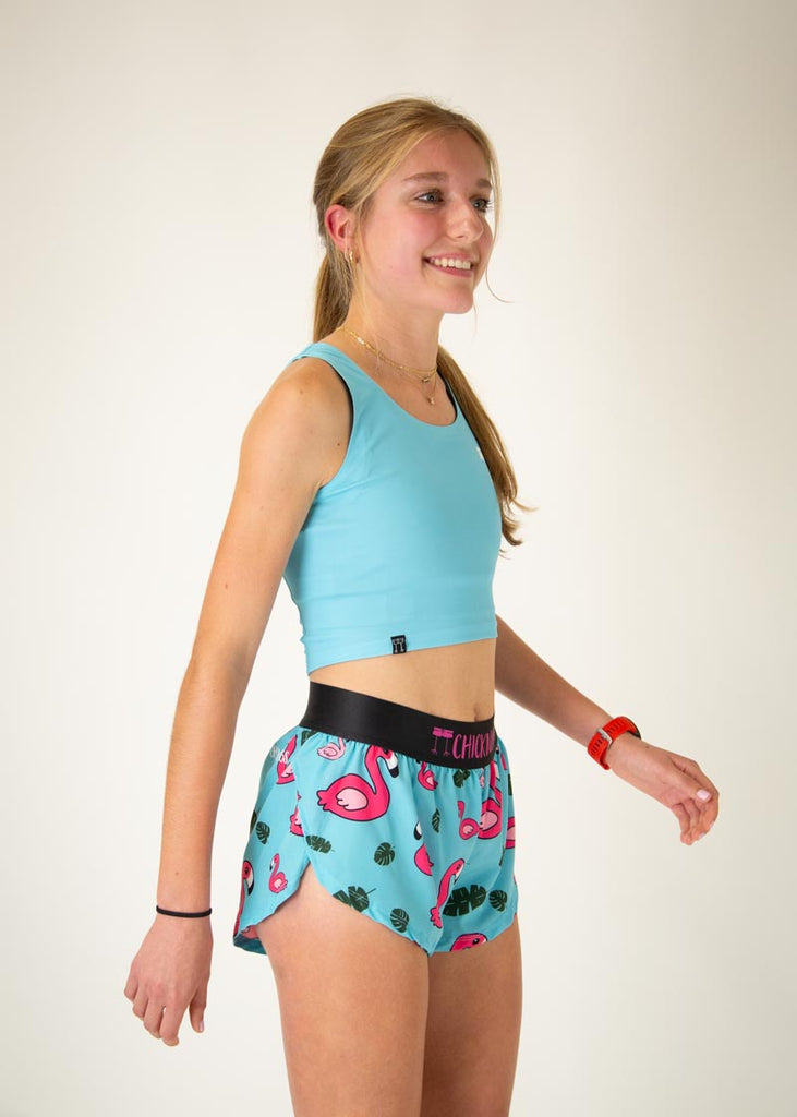 The model is a runner who is wearing Chicknlegs women's pwr crop top in the sky blue design, facing front. The model is matching the pwr crop with the women's 1.5" split running shorts in the blue flamingo design. The model is facing right.