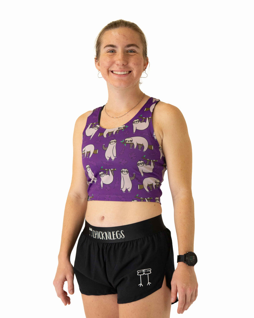 Runner is wearing ChicknLegs in the women's power crop top in the sloths design. The material is lightweight and the print has sloths with branches and leaves with a solid purple background.
