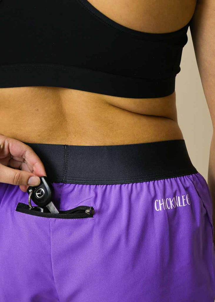 Model is wearing Chicknlegs women's 1.5 inch split running shorts in the purple design close up shot of model putting keys into back pocket.